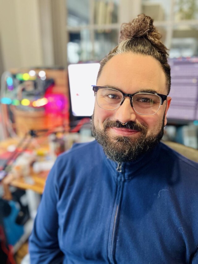 Ryan Tropf — smiling, with a top-knot, beard, and glasses, wearing a blue zip-up